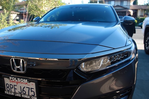 A sleek, shiny gray Honda car is parked in a residential driveway. The front view shows the vehicle's headlights and grille, with a visible Folsom Lake Honda license plate. In the background are trees and the facade of a house, along with a person walking nearby.