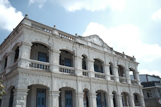A historic two-story building with ornate architectural details, featuring arches, columns, and decorative moldings. The structure is painted in a light color, and the windows have blue shutters. The sky is bright with scattered clouds, adding to the serene atmosphere.