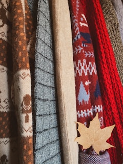 A collection of knitted sweaters in various textures and patterns, with earthy and autumnal colors. There is a maple leaf tucked into one of the sweaters, adding to the autumnal theme.