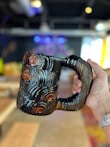 A hand is holding a unique, ceramic mug with a textured, grooved pattern that resembles tree rings. The mug has a glossy finish with dark and light brown tones. The background is an indoor space with blurred elements, including furniture and colorful lights, making the mug the primary focus.