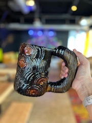A hand is holding a unique, ceramic mug with a textured, grooved pattern that resembles tree rings. The mug has a glossy finish with dark and light brown tones. The background is an indoor space with blurred elements, including furniture and colorful lights, making the mug the primary focus.