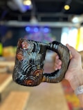 A hand is holding a unique, ceramic mug with a textured, grooved pattern that resembles tree rings. The mug has a glossy finish with dark and light brown tones. The background is an indoor space with blurred elements, including furniture and colorful lights, making the mug the primary focus.