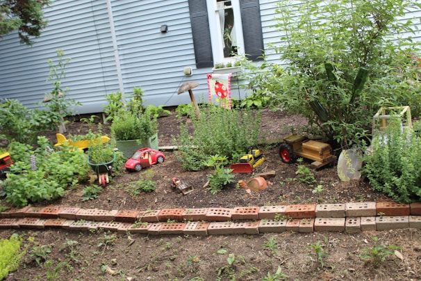 A small garden area with a variety of green plants and herbs is bordered by a row of bricks. Several toy vehicles, including a red car and a truck, are scattered around the garden. A small banner with a sunflower design hangs from a wooden post. In the background, a blue house with black shutters and a window with flower boxes is visible.