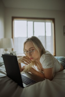 A person with long hair and glasses is lying on a bed while focusing on a laptop. The room appears softly lit with a window in the background letting in natural light. A lamp and some curtains are visible, suggesting a cozy, relaxed environment.