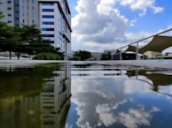 A modern urban landscape featuring a large office building with blue-tinted windows reflects in a calm water surface. Fluffy clouds in a bright blue sky are mirrored in the water. Surrounding the building are lush green trees, and there are modern architectural structures with curved roofs visible on the right.