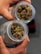 Two hands hold glass jars filled with cannabis buds. The buds are dense and have a mix of green and brown hues. The jars have a frosted surface with a pattern. The background is blurred, emphasizing the contents of the jars.