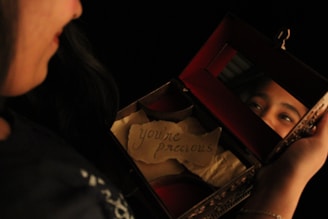A person is gazing into a small, open box with a mirror inside that reflects part of their face. The box contains a piece of paper with the handwritten message 'you're precious'. The lighting is warm and intimate, highlighting the reflection and the inscription.