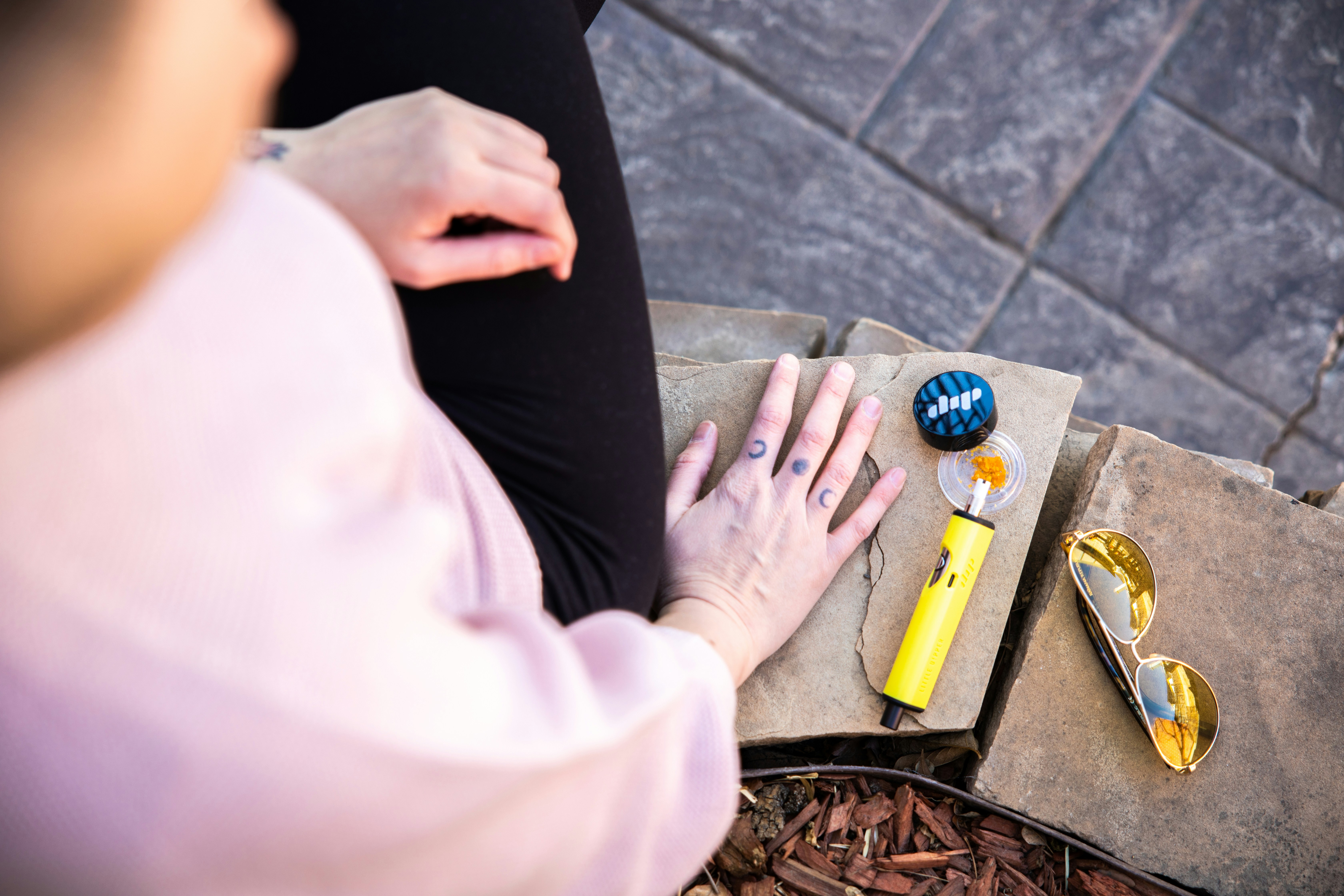A woman in a pink sweater sits on a stone wall beside a yellow Dip Devices Little Dipper dab pen, yellow sunglasses and cannabis dabs. Get yours at https://dipdevices.com/