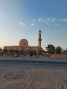 A mosque with a large dome and tall minaret against a clear blue sky. Palm trees line the building along a quiet road with tire tracks in the sand.