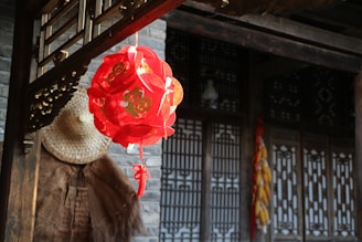 A red lantern with Chinese characters hanging under a wooden structure, surrounded by traditional architecture with wooden lattice windows. A straw hat and a bunch of dried corn cobs are visible, enhancing the rustic and cultural atmosphere.
