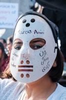 A person is wearing a white mask with red speckles, featuring cutout eyes and mouth. Written on the mask are the phrases 'Women Life Freedom' and '#MansaAmini', indicating a possible protest or activist context. The background shows partially blurred text and other people in an outdoor setting.