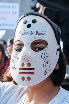 A person is wearing a white mask with red speckles, featuring cutout eyes and mouth. Written on the mask are the phrases 'Women Life Freedom' and '#MansaAmini', indicating a possible protest or activist context. The background shows partially blurred text and other people in an outdoor setting.