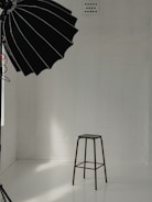 A minimalist studio setup with a tall stool positioned against a white brick wall. A large black photography umbrella is partly visible, casting light onto the scene. The floor is smooth and reflective, creating a clean and bright atmosphere.