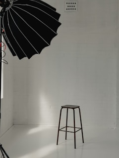 A minimalist studio setup with a tall stool positioned against a white brick wall. A large black photography umbrella is partly visible, casting light onto the scene. The floor is smooth and reflective, creating a clean and bright atmosphere.