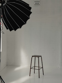 A minimalist studio setup with a tall stool positioned against a white brick wall. A large black photography umbrella is partly visible, casting light onto the scene. The floor is smooth and reflective, creating a clean and bright atmosphere.