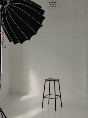 A minimalist studio setup with a tall stool positioned against a white brick wall. A large black photography umbrella is partly visible, casting light onto the scene. The floor is smooth and reflective, creating a clean and bright atmosphere.