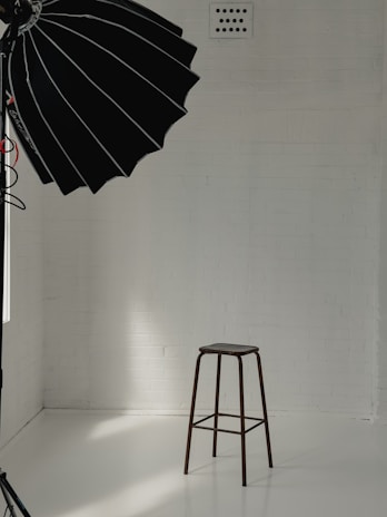 A minimalist studio setup with a tall stool positioned against a white brick wall. A large black photography umbrella is partly visible, casting light onto the scene. The floor is smooth and reflective, creating a clean and bright atmosphere.