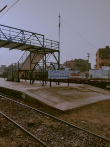 A railway platform with a sign reading 'Hyderabad JN'. The platform is empty, with a metal pedestrian bridge crossing above. The surrounding area includes industrial buildings and utility poles with visible wires. The ground appears dusty, and the sky has a hazy, muted tone.