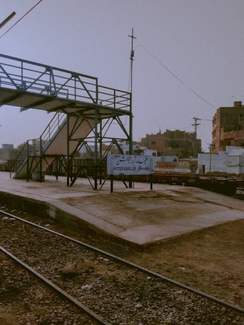 A railway platform with a sign reading 'Hyderabad JN'. The platform is empty, with a metal pedestrian bridge crossing above. The surrounding area includes industrial buildings and utility poles with visible wires. The ground appears dusty, and the sky has a hazy, muted tone.