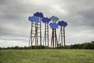 Several tall structures with cloud-shaped tops in various shades of blue are placed in an open grassy field. The cloudy sky above adds to the surreal and artistic nature of the installation. Trees are visible in the background, along the horizon.