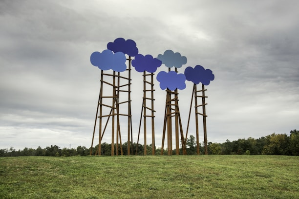 Several tall structures with cloud-shaped tops in various shades of blue are placed in an open grassy field. The cloudy sky above adds to the surreal and artistic nature of the installation. Trees are visible in the background, along the horizon.