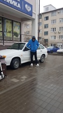 A person in a blue jacket and black pants stands beside a white car parked on wet pavement. The individual has their hands in their pockets and is facing downward. Behind them, there is a building with commercial signs and several windows.