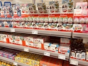 Shelves stocked with various kinds of Kinder Surprise chocolate eggs and boxes. The display includes colorful packaging with themed designs featuring animals and popular characters. Price tags are visible, indicating various prices for different products.