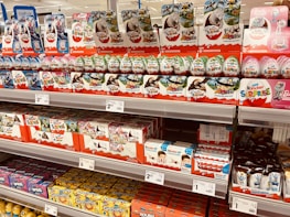 Shelves stocked with various kinds of Kinder Surprise chocolate eggs and boxes. The display includes colorful packaging with themed designs featuring animals and popular characters. Price tags are visible, indicating various prices for different products.