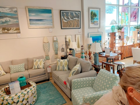 A cozy living room setup in a showroom features two gray couches with patterned geometric pillows. A matching patterned coffee table holds a decorative tray. The room is adorned with nautical-themed artwork and decorative elements like pendant lights and wooden wall art shaped like fish. Large windows allow natural light to flood in, and multiple lamps are displayed on wooden side tables.