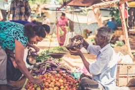 A lively outdoor market scene with a vendor weighing produce using a traditional balance scale. A woman examines fresh tomatoes and other vegetables arranged on a table. Bright sunlight illuminates the setting, highlighting the fruits and vegetables as well as the people present.