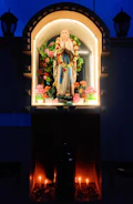 A statue of a religious figure, adorned with garlands, is housed within a niche. The surrounding area is decorated with vibrant flowers, and candles are lit at the base, creating a warm glow against the dark backdrop.