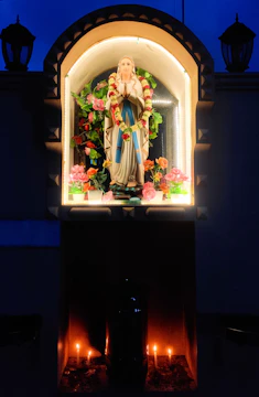 A statue of a religious figure, adorned with garlands, is housed within a niche. The surrounding area is decorated with vibrant flowers, and candles are lit at the base, creating a warm glow against the dark backdrop.