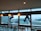 Two workers are suspended outside a large glass window, engaged in cleaning the exterior of a modern high-rise building. The workers are using ropes for support, wearing safety harnesses and gear. Inside, there is a row of empty chairs lined up against a bar-style table. The glass allows a view of other tall buildings with a slightly cloudy sky in the background.