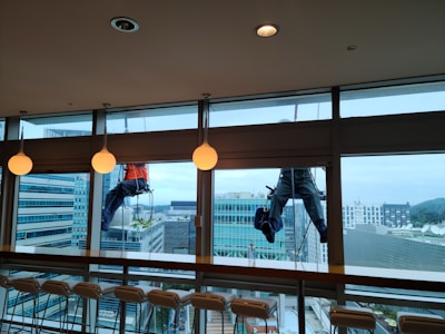 Two workers are suspended outside a large glass window, engaged in cleaning the exterior of a modern high-rise building. The workers are using ropes for support, wearing safety harnesses and gear. Inside, there is a row of empty chairs lined up against a bar-style table. The glass allows a view of other tall buildings with a slightly cloudy sky in the background.