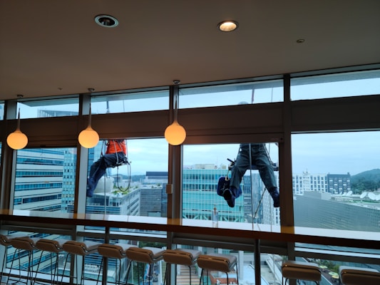 Two workers are suspended outside a large glass window, engaged in cleaning the exterior of a modern high-rise building. The workers are using ropes for support, wearing safety harnesses and gear. Inside, there is a row of empty chairs lined up against a bar-style table. The glass allows a view of other tall buildings with a slightly cloudy sky in the background.