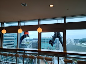Two workers are suspended outside a large glass window, engaged in cleaning the exterior of a modern high-rise building. The workers are using ropes for support, wearing safety harnesses and gear. Inside, there is a row of empty chairs lined up against a bar-style table. The glass allows a view of other tall buildings with a slightly cloudy sky in the background.