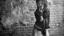 A woman with dark hair is wearing a leather jacket and posing energetically against a textured brick wall. Her facial expression is enthusiastic, with her mouth open as if shouting or singing. Her hands are positioned in a rock and roll gesture, and the setting has an edgy, urban feel with the wall showing signs of wear.