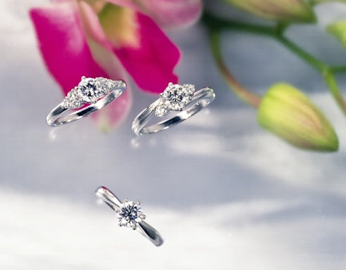 Three diamond rings are elegantly arranged against a soft, blurred floral background. The rings are silver with centered sparkling diamonds, with some featuring additional smaller stones on the sides. The out-of-focus flowers in the background add a subtle touch of pink and green hues, enhancing the image's romantic and luxurious feel.