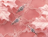 Three elegant rings featuring sparkling diamonds are artistically arranged against a delicate pink floral lace background. The diamonds catch the light, adding a touch of glamour and sophistication.