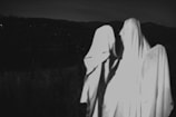 Three figures covered in white sheets appear to resemble ghosts standing in a dark outdoor setting. The background includes silhouetted trees and a hilly landscape under dim lighting.