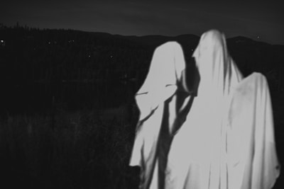 Three figures covered in white sheets appear to resemble ghosts standing in a dark outdoor setting. The background includes silhouetted trees and a hilly landscape under dim lighting.