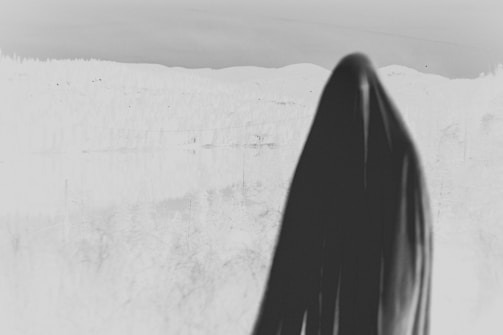 A dark, cloaked figure stands against a stark, negative space, creating a haunting and mysterious ambiance. The black garment contrasts sharply with the lighter, washed-out background that resembles a desolate or eerie landscape.