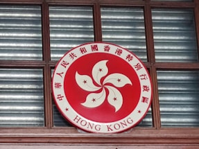 A circular emblem is displayed, featuring a red background with a stylized white flower in the center and five stars. Chinese characters surround the flower, along with the words 'HONG KONG' at the bottom. The emblem is mounted on a wooden window frame with glass panes behind it.