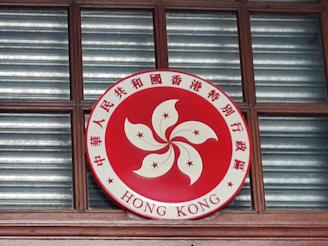 A circular emblem is displayed, featuring a red background with a stylized white flower in the center and five stars. Chinese characters surround the flower, along with the words 'HONG KONG' at the bottom. The emblem is mounted on a wooden window frame with glass panes behind it.