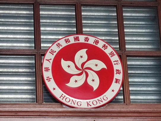 A circular emblem is displayed, featuring a red background with a stylized white flower in the center and five stars. Chinese characters surround the flower, along with the words 'HONG KONG' at the bottom. The emblem is mounted on a wooden window frame with glass panes behind it.