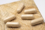 Six beige capsules are spread out on a coarse, wooden surface with a finely textured background.
