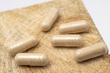 Six beige capsules are spread out on a coarse, wooden surface with a finely textured background.