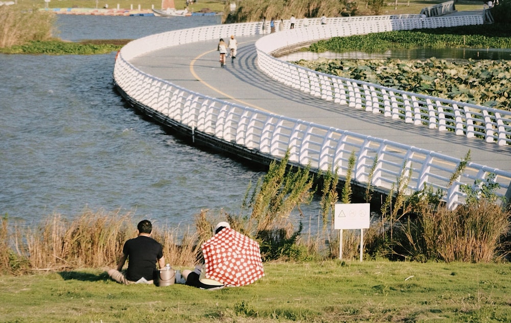a couple of people sitting on a grassy hill by a body of water