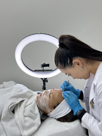 A beauty technician carefully performs a skincare procedure on a woman, who is lying down with her hair pulled back and face covered in a transparent mask. The technician is wearing gloves and holding a precision tool, working under a bright ring light.