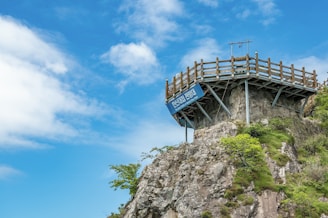 A wooden viewing platform is perched atop a rocky cliff, surrounded by lush green vegetation under a bright blue sky with scattered white clouds. A sign is attached to the platform.